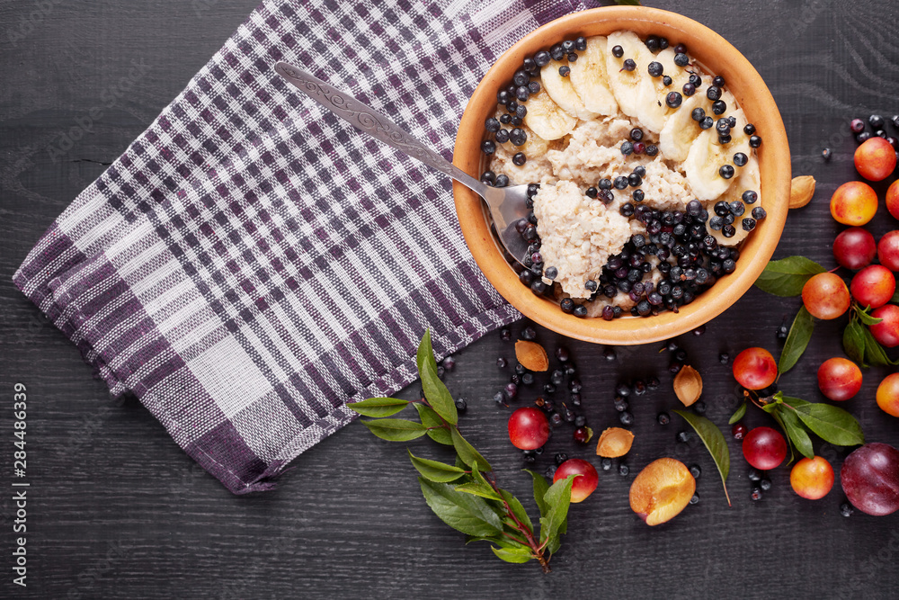 Picture of porridge with blueberries and banana slices in white plate on dark concrete background. Decoration of green leaves, aliches, plums and checkered cotton towels. Healthy breakfast concept. Picture of porridge with blueberries and banana slices in white plate on dark concrete background. Decoration of green leaves, aliches, plums and checkered cotton towels. Healthy breakfast concept.