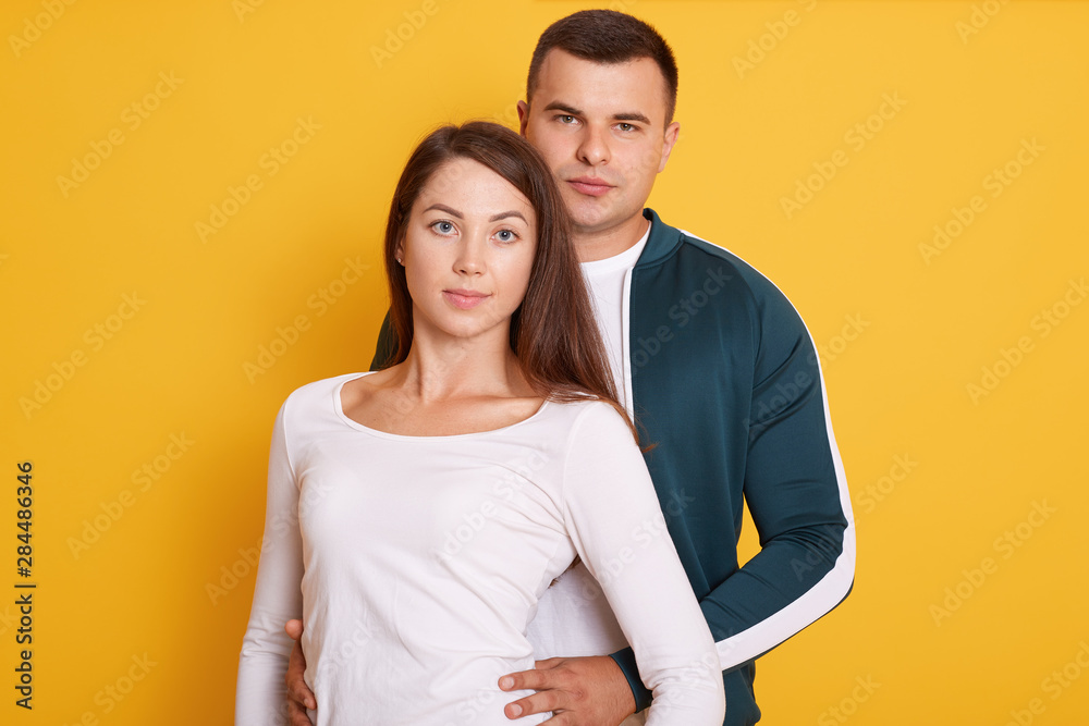 Closeup portrait of beautiful young couple isolated on yellow background. Attractive man and woman looking at camera, guy in casual shirt hugs her girlfriend, female wearing white shirt. Love concept. Closeup portrait of beautiful young couple isolated on yellow background. Attractive man and woman looking at camera, guy in casual shirt hugs her girlfriend, female wearing white shirt. Love concept.