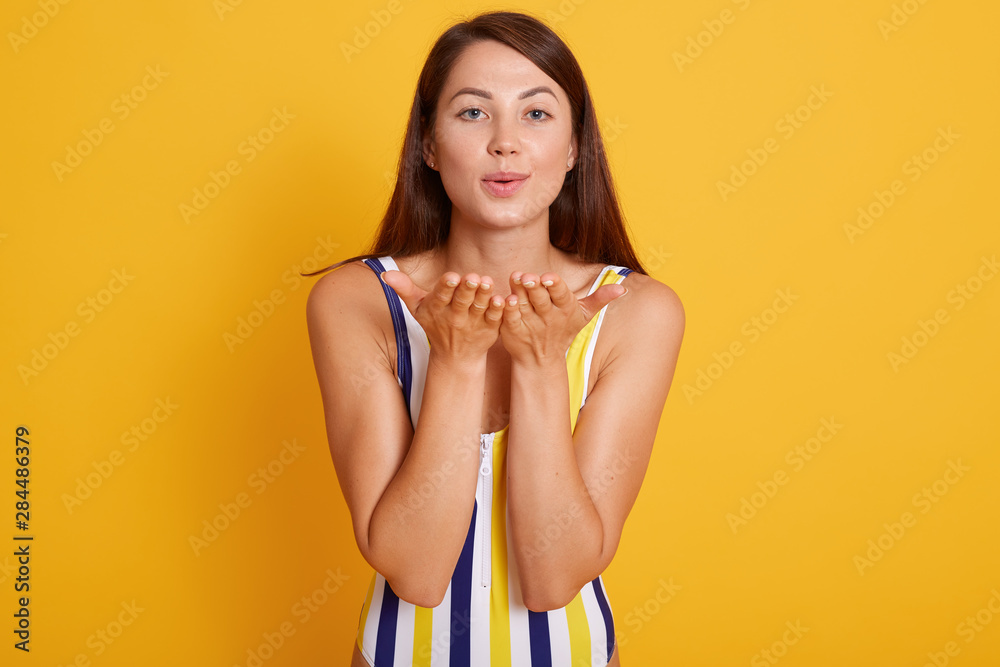 Naklejka premium Horizontal shot of pleasant looking female with healthy skin, wearing swimsuit, making air kiss gesture, flirts with handsome guy at seaside, isolated over yellow studio background. People concept.