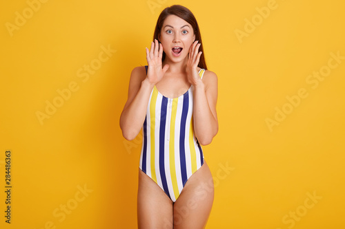 Photo of shocked young woman in srtiped swimsuit looking directly at camera, isolated over yellow studio wall, keeps hands near mouth, crying something, posing with widely opened eyes. People concept. photo
