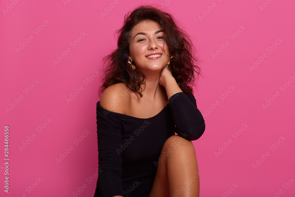 Beautiful girl with dark wavy hair, sitting isolated over pink studio background, keeps hand on neck, posing with bare shoulders and wearing fashionable black shirt, looking smiling directly at camera Beautiful girl with dark wavy hair, sitting isolated over pink studio background, keeps hand on neck, posing with bare shoulders and wearing fashionable black shirt, looking smiling directly at camera