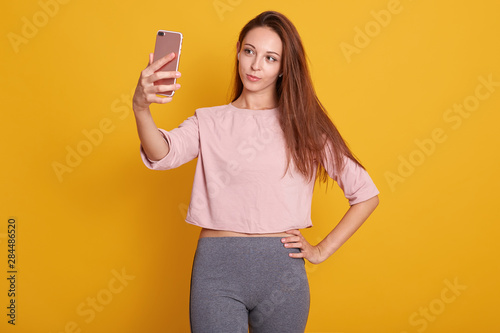Studio shot of beautiful brown haired woman with straight hair in gray trousers and rose shirt taking selfie with mobile phone, holds her device in hand, posing isolated over yellow background. photo