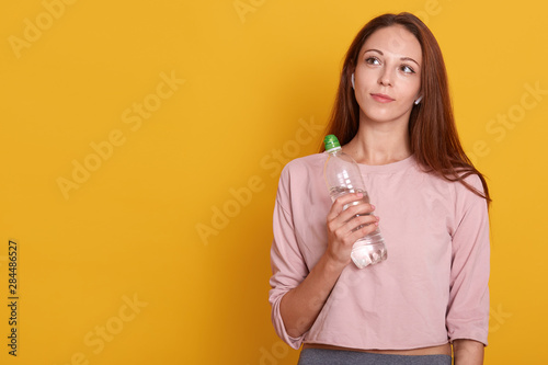 Young athletic woman wearing wireless headphones and rose sportswear holds bottle of water in hands, looking up, ready for running, isolated over yellow wall, copy space for promotion or advertismant. photo