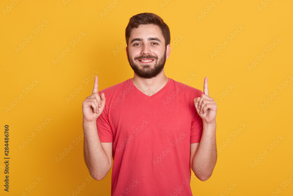 Image of young happy man with good mood, posing isolated over yellow background, pointing up with his index fingers, looking smiling directly at camera. Copy space for advertisment or promotion. Image of young happy man with good mood, posing isolated over yellow background, pointing up with his index fingers, looking smiling directly at camera. Copy space for advertisment or promotion.