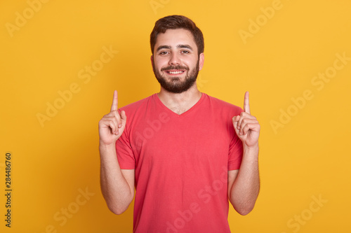 Image of young happy man with good mood, posing isolated over yellow background, pointing up with his index fingers, looking smiling directly at camera. Copy space for advertisment or promotion.
