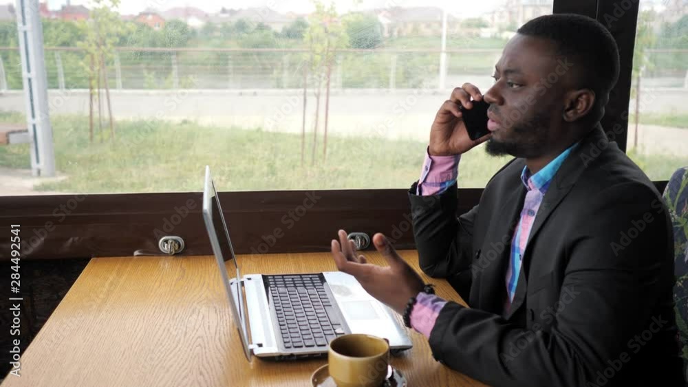 Afro american businessman working on laptop and talking mobile phone in cafe. He has a cup of coffee. Black man calling mobile phone and looking documents on computer. He wears shirt and suit jacket. Afro american businessman working on laptop and talking mobile phone in cafe. He has a cup of coffee. Black man calling mobile phone and looking documents on computer. He wears shirt and suit jacket.