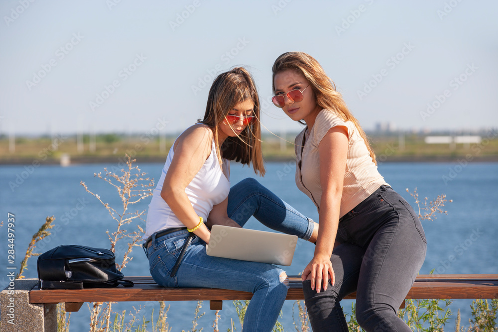 Two beautiful young caucasian girls looking amazed to a laptop while sitting outside in the park on a bench.