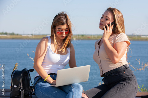 Two smiling young girls using laptop at the park, laughing, chatting