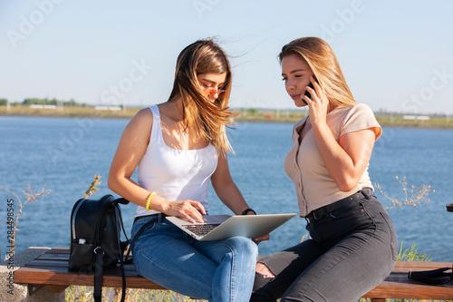Two smiling young girls using laptop at the park, laughing, chatting