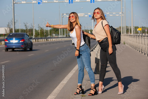 Two beautiful young women are hitchhiking on the road and try to find someone to take them to their hotel.