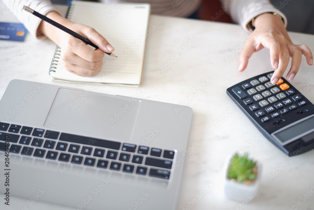 Closed up and focus on hand of young woman using a calculator and other hand using pencil take a note in the book and have notebook on the table at cafe or co working space. co working space concept.