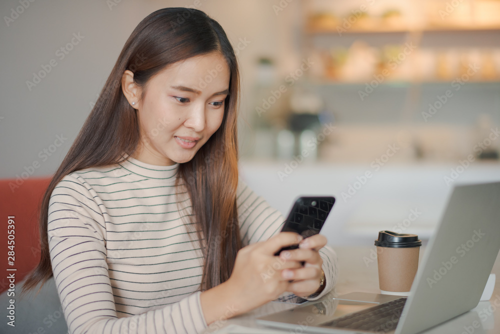 Young beautiful Asian woman working with smartphone and notebook laptop. She is holding a smartphone on hand. Woman smiling and felling happy which looking a phone at cafe. co working space concept.