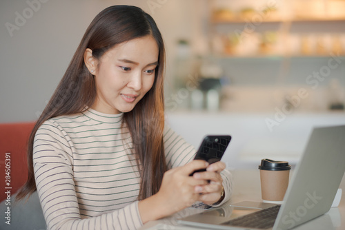 Young beautiful Asian woman working with smartphone and notebook laptop. She is holding a smartphone on hand. Woman smiling and felling happy which looking a phone at cafe. co working space concept.