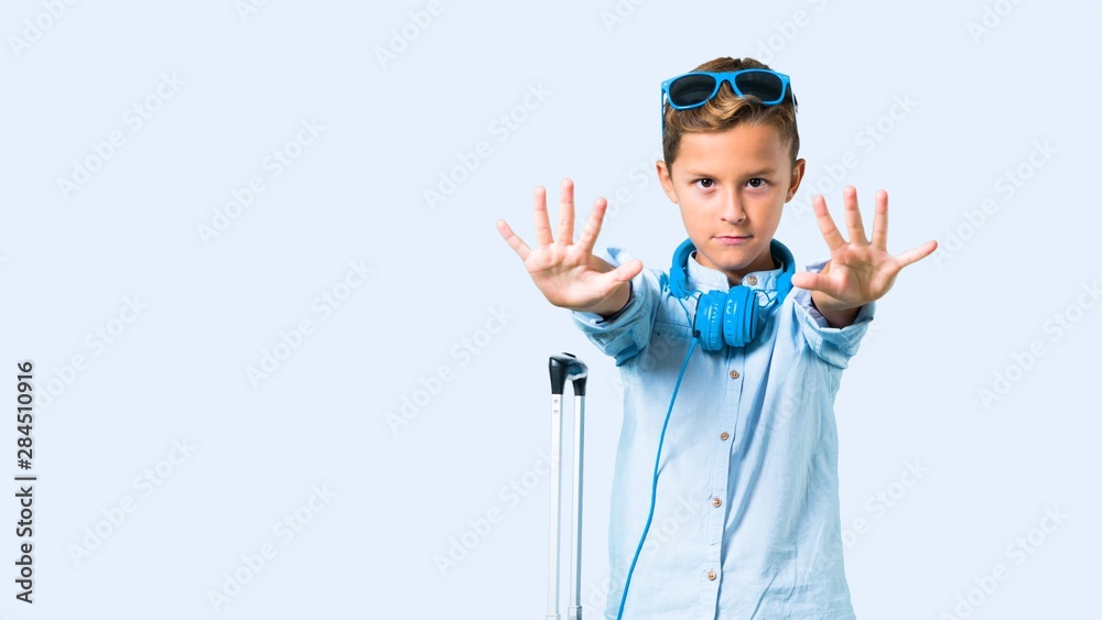 Fototapeta premium Kid with sunglasses and headphones traveling with his suitcase making stop gesture with her hand on blue background