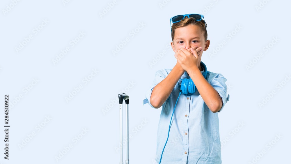Kid with sunglasses and headphones traveling with his suitcase covering mouth with hands on blue background