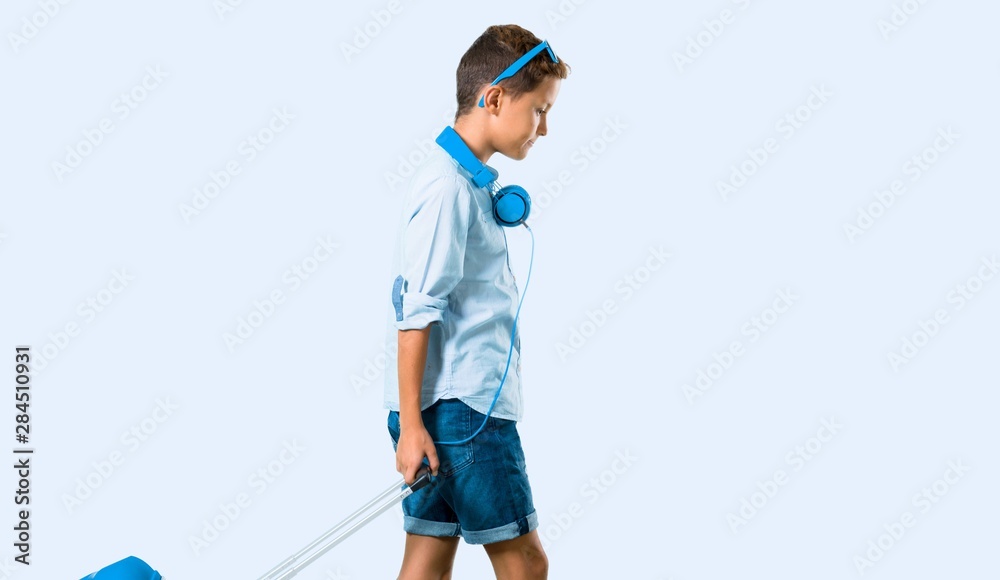 Boy with sunglasses and headphones traveling with his suitcase on blue background Boy with sunglasses and headphones traveling with his suitcase on blue background