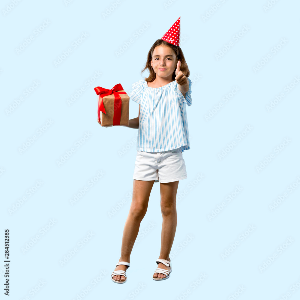 Full body of Little girl at a birthday party holding a gift giving a thumbs up gesture and smiling on blue background Full body of Little girl at a birthday party holding a gift giving a thumbs up gesture and smiling on blue background