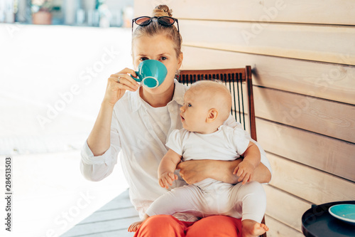 Caucasian young woman sits in a deck chair at the table, drinks coffee and holds the baby in her arms. Summer day in the city. Lifestyle portrait of mother with baby. photo