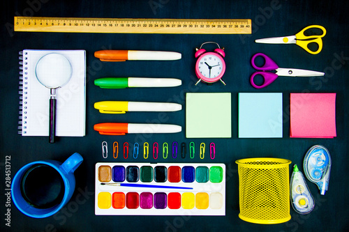 Colorful office supplies neatly geometrically evenly laid out on blackboard background. Markers, scissors, watercolors, cup, alarm clock, paper clips on canvas. Preparing perfectionist for study.