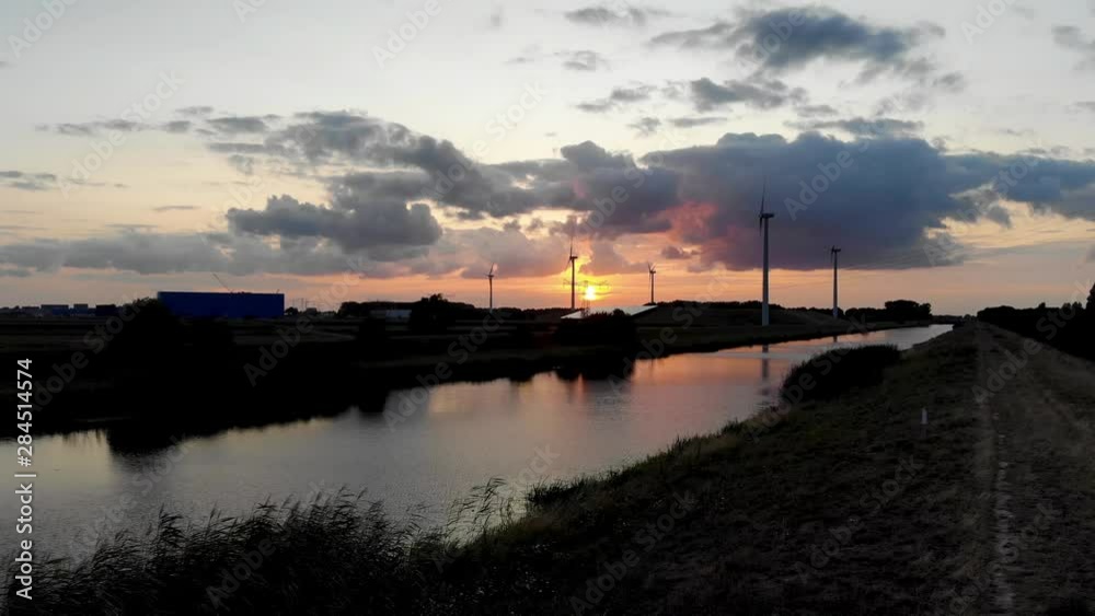 Aerial low angle dolly of Windmills in countryside at sunset. Drone fly over river with reflection of colorful sky, green fields. Rural landscape with farm and factory in nature. Aerial low angle dolly of Windmills in countryside at sunset. Drone fly over river with reflection of colorful sky, green fields. Rural landscape with farm and factory in nature.