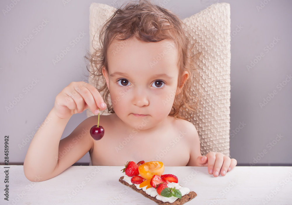 Little cute girl holding cherry in a hand and eating crisp bread with creme cheese, fruit and berries. Little cute girl holding cherry in a hand and eating crisp bread with creme cheese, fruit and berries.