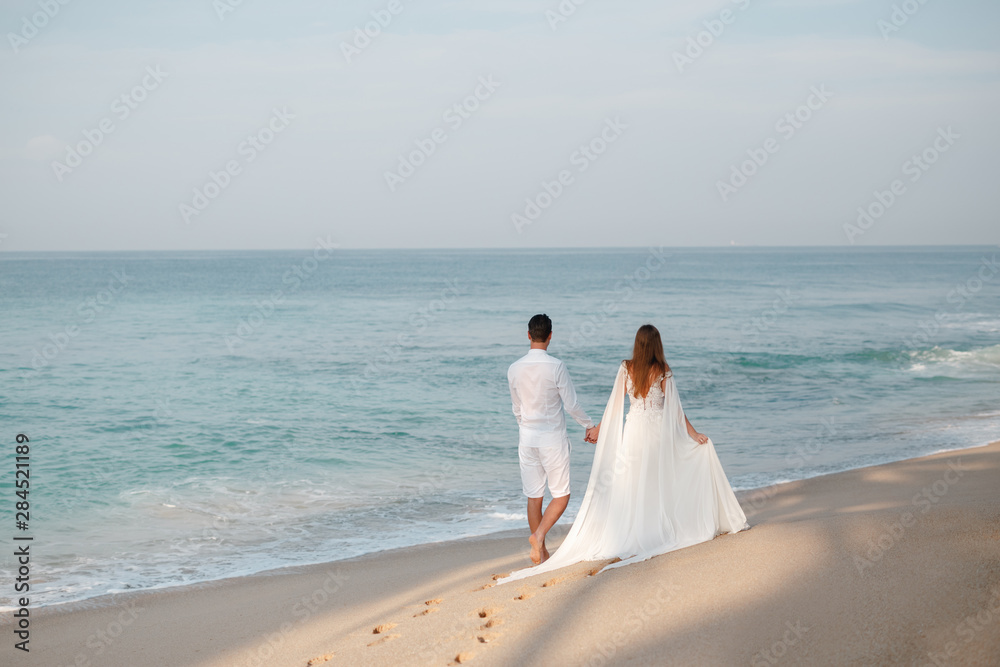 Romantic young couple walking together on beautiful exotic tropical beach, enjoy their honeymoon at the ocean. back view, unrecognizable Romantic young couple walking together on beautiful exotic tropical beach, enjoy their honeymoon at the ocean. back view, unrecognizable