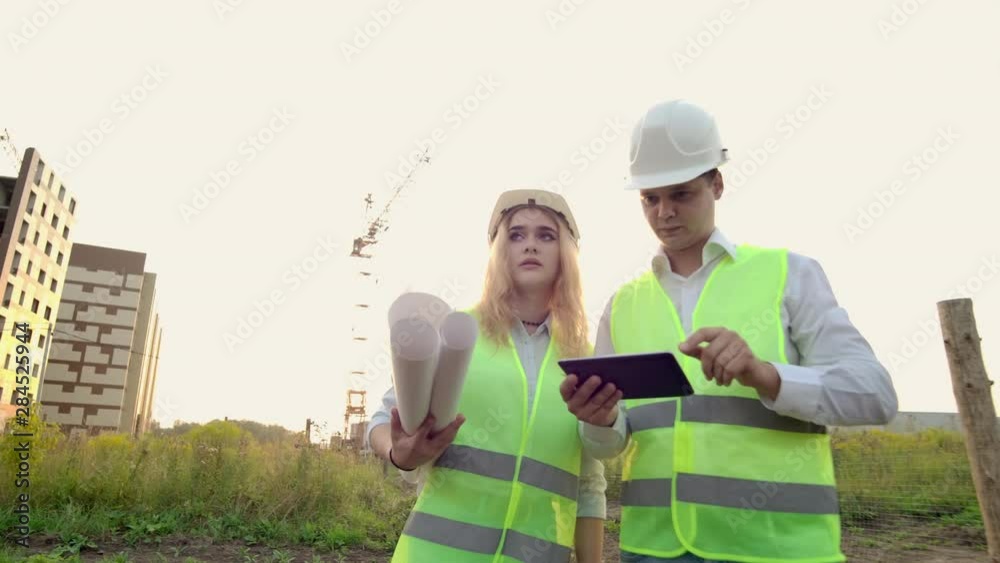 Adult engineer man and architect woman uses a tablet in operation. Writes a message or checks a drawing. Against background is building.