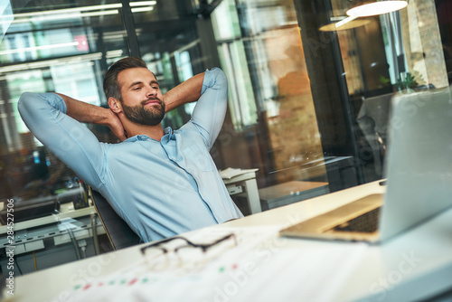 Work done Satisfied young bearded businessman leaning back with hands behind head and smiling while sitting in the modern office photo