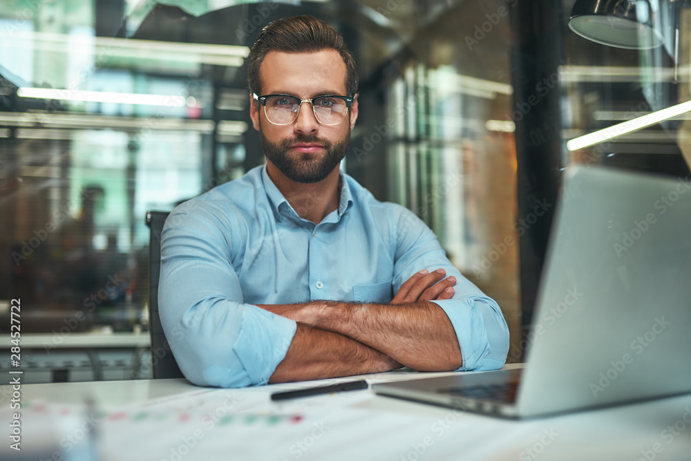 Confidence. Young bearded businessman in eyeglasses and formal wear looking at camera and keeping arms crossed while sitting in modern office - obrazy, fototapety, plakaty 