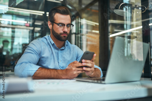 Important call. Portrait of young focused businessman in eyeglasses and formal wear looking at his smartphone while sitting in the office