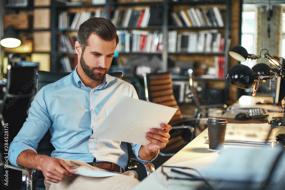 Let me see. Young and handsome bearded man in formal wear examining documents while sitting in his modern office. Working place Let me see. Young and handsome bearded man in formal wear examining documents while sitting in his modern office. Working place