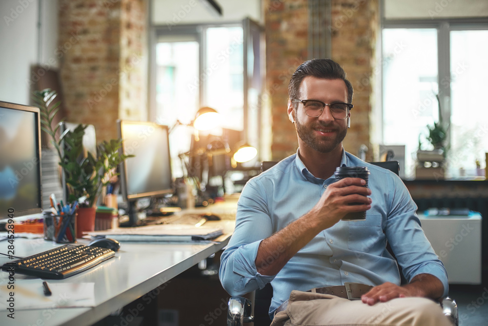 © Friends Stock - I can not work without coffee. Portrait of young bearded man in eyeglasses and headphones holding cup of coffee and smiling at camera while sitting in the modern office © Friends Stock - I can not work without coffee. Portrait of young bearded man in eyeglasses and headphones holding cup of coffee and smiling at camera while sitting in the modern office