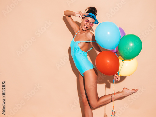 Young beautiful sexy hipster woman in sunglasses and transparent visor cap.Trendy girl in summer swimwear bathing suit. Positive female with colorful air balloons.Ready for party