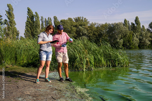 ecologists on river bank examine sample of green algae and enter info on tablet