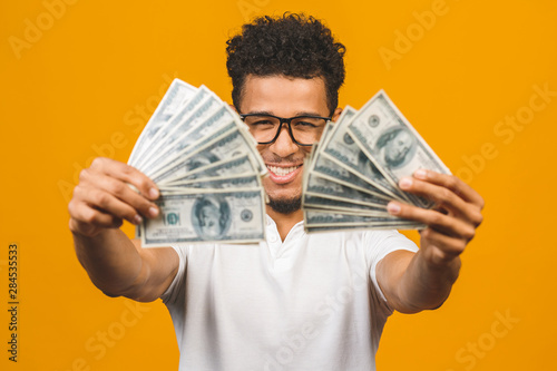 Winner! Young rich african american man in casual t-shirt holding money dollar bills with surprise isolated over yellow wall.