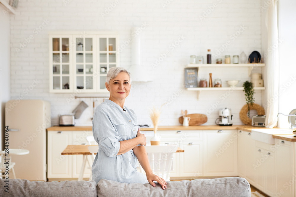 Horizontal image of gorgeous joyful elderly sixty year old housewife having rest after cleaning all rooms, having cheerful facial expression, standing in living room with kitchen in background Horizontal image of gorgeous joyful elderly sixty year old housewife having rest after cleaning all rooms, having cheerful facial expression, standing in living room with kitchen in background
