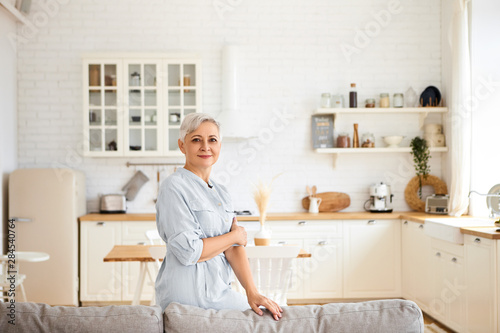 Horizontal image of gorgeous joyful elderly sixty year old housewife having rest after cleaning all rooms, having cheerful facial expression, standing in living room with kitchen in background