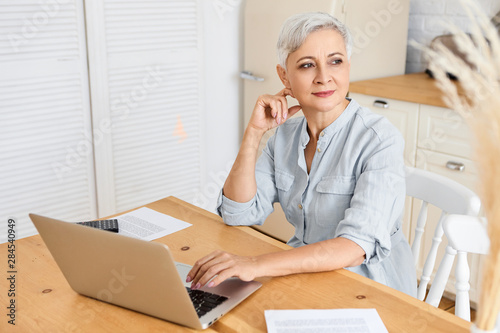 Commincation, technology, job, age and retirement concept. Concentrated serious self employed female pensioner sitting in kitchen counter, keyboarding on laptop, earning living via internet