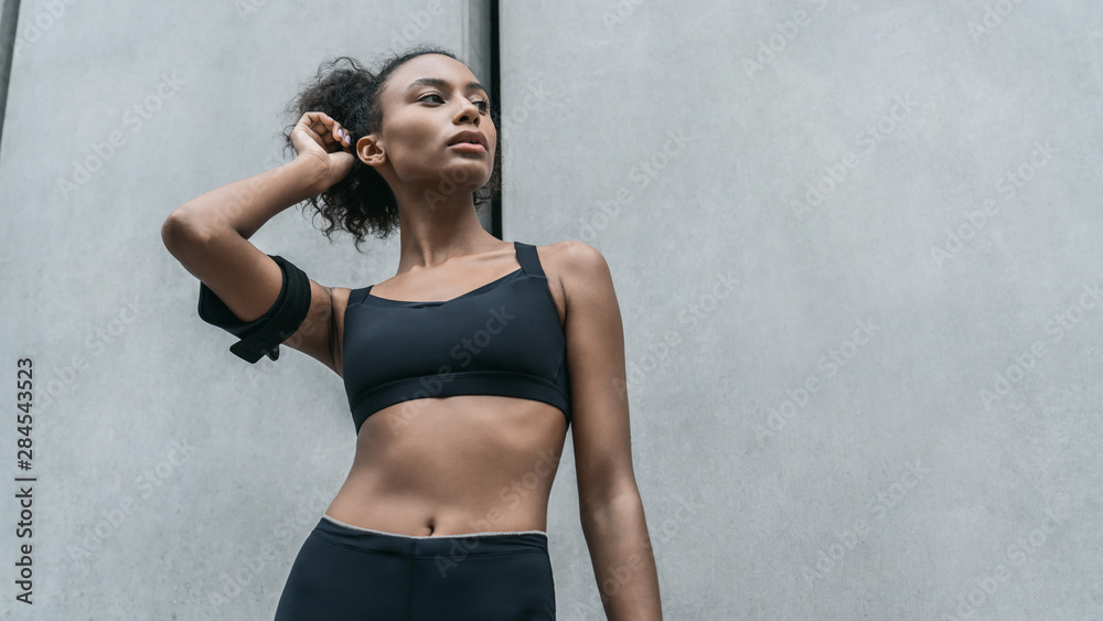 Fototapeta premium Female young athlete taking a break after training outdoors standing against grey wall