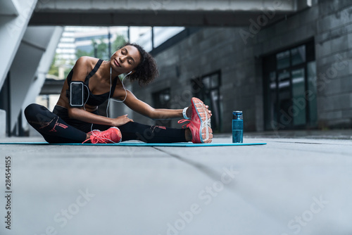 Young female with earphones exercising and stretching outdoors on fitness mat