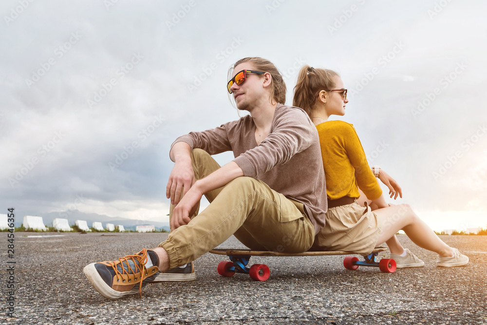 An attractive young couple in sunglasses sit back to back on their longboard in a suburban parking lot against a gray sky. The concept of a young family generation millennials style and leisure