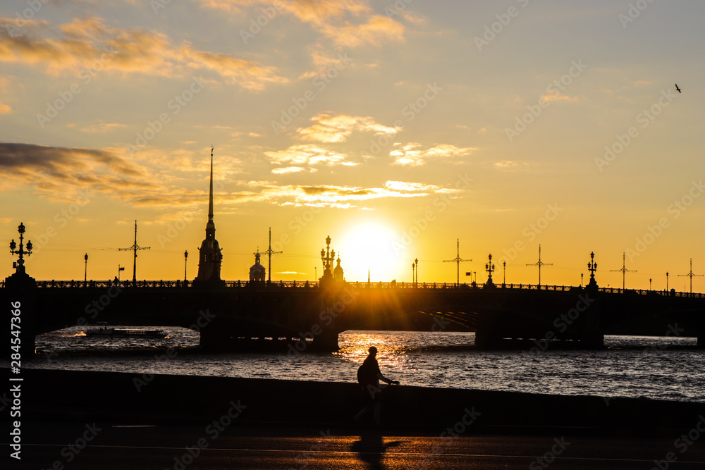 View of the Neva River, Trinity Bridge, Peter and Paul Fortress. Saint-Petersburg, Russia. Summer sunset on the embankment. Silhouette of a man walking along the embankment at sunset. View of the Neva River, Trinity Bridge, Peter and Paul Fortress. Saint-Petersburg, Russia. Summer sunset on the embankment. Silhouette of a man walking along the embankment at sunset.