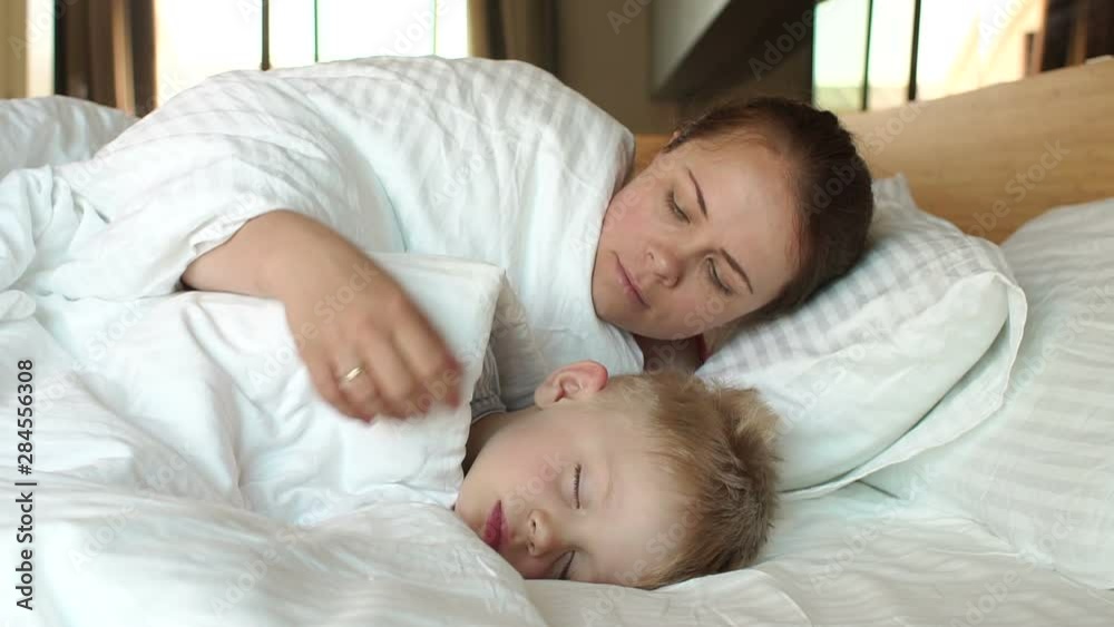 A gentle caring mother hugs and strokes the head of her sleeping son in bed. Mom and baby sleep in bed in the morning, mom covers her baby with a blanket. Close up, slow motion. A gentle caring mother hugs and strokes the head of her sleeping son in bed. Mom and baby sleep in bed in the morning, mom covers her baby with a blanket. Close up, slow motion.