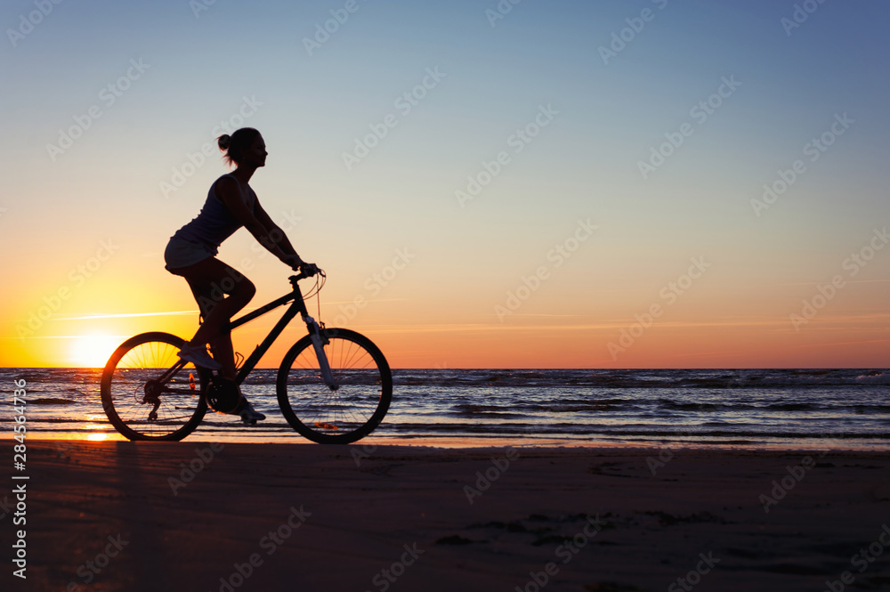 Fototapeta premium Silhouette of sporty woman riding bicycle on multicolored sunset background