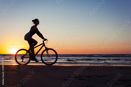 Silhouette of sporty woman riding bicycle on multicolored sunset background