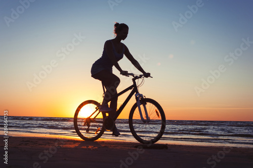 Silhouette of sporty woman riding bicycle on multicolored sunset background