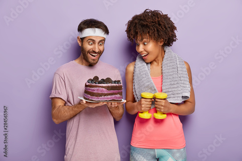 Joyous female and male stare with happiness and temptation at delicious cake, being hungry after exhausted workout, avoid eating sweet desserts with much calories, exercise with dumbbells in gym
