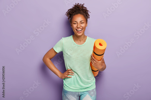 Overjoyed healthy dark skinned female athlete keeps hand on hip, holds rolled up fitness mat, being in good physical shape, has sport training every day, wears t shirt and leggings. People, yoga photo
