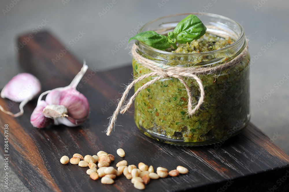 Pesto green sauce in a glass jar. On a wooden board next to a jar of garlic and pine nuts. Close-up. Gray background. Pesto green sauce in a glass jar. On a wooden board next to a jar of garlic and pine nuts. Close-up. Gray background.
