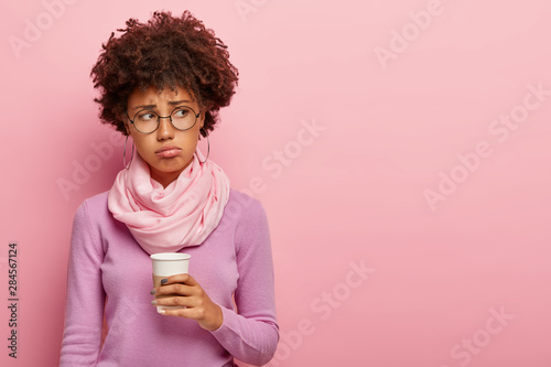 Studio shot of displeased dark skinned female drinks fresh coffee as morning refreshment, looks away in dissatisfaction, dressed in purple jumper and scarf on neck poses over rosy wall with copy space